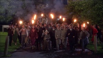 Movie still from “Reefer Madness: The Movie Musical” (2005), directed by Andy Fickman – A group of people in costumes are holding torches in the street; Extreme Wide shot, High angle