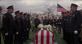 Movie still from “Remo Williams: The Adventure Begins” (1985), directed by Guy Hamilton – A group of men standing next to an american flag draped casket; Wide shot, High angle