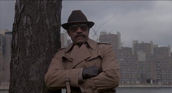 Movie still from “Remo Williams: The Adventure Begins” (1985), directed by Guy Hamilton – A man in a trench coat and a hat standing next to a tree; Medium shot, Low angle