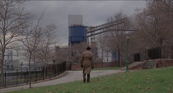Movie still from “Remo Williams: The Adventure Begins” (1985), directed by Guy Hamilton – A man in a trench coat walking on a path near trees; Extreme Wide shot, High angle