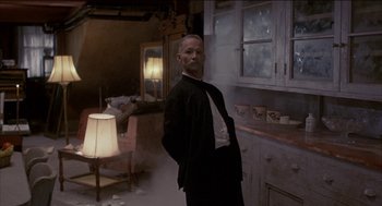 Movie still from “Remo Williams: The Adventure Begins” (1985), directed by Guy Hamilton – A man standing in a room with a lamp on a table; Medium shot, Low angle