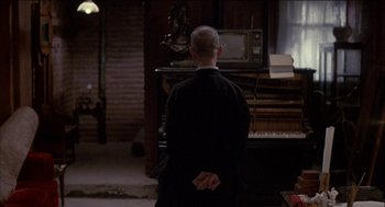 Movie still from “Remo Williams: The Adventure Begins” (1985), directed by Guy Hamilton – A man standing in front of an old fashioned piano; Wide shot, Over the shoulder angle