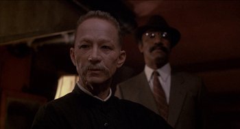 Movie still from “Remo Williams: The Adventure Begins” (1985), directed by Guy Hamilton – A man in a suit and tie next to another man in a suit and tie; Close Up shot, Low angle