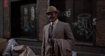 Movie still from “Remo Williams: The Adventure Begins” (1985), directed by Guy Hamilton – A man in a suit and hat is holding a bag; Medium shot, Low angle