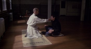 Movie still from “Remo Williams: The Adventure Begins” (1985), directed by Guy Hamilton – A man sitting on the ground with a man sitting on the floor; Wide shot, High angle