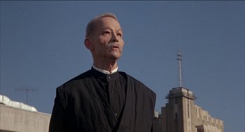 Movie still from “Remo Williams: The Adventure Begins” (1985), directed by Guy Hamilton – An older man wearing a black shirt and a white shirt; Medium shot, Low angle