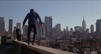 Movie still from “Remo Williams: The Adventure Begins” (1985), directed by Guy Hamilton – A man riding a skateboard on top of a building; Wide shot, Low angle