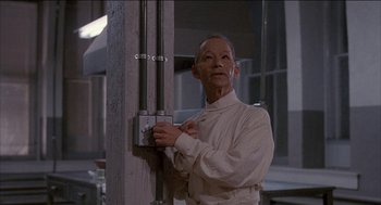 Movie still from “Remo Williams: The Adventure Begins” (1985), directed by Guy Hamilton – An older man in a white shirt is leaning against a pole; Medium shot, Low angle