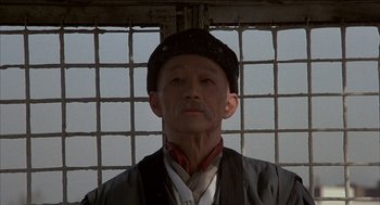 Movie still from “Remo Williams: The Adventure Begins” (1985), directed by Guy Hamilton – An older man wearing a black hat and a black jacket; Close Up shot, Low angle