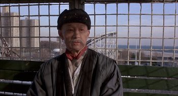 Movie still from “Remo Williams: The Adventure Begins” (1985), directed by Guy Hamilton – An older man wearing a hat standing in front of a bridge; Medium shot, Low angle
