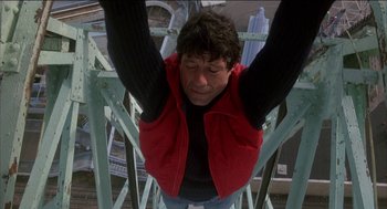 Movie still from “Remo Williams: The Adventure Begins” (1985), directed by Guy Hamilton – A man hanging from a metal rail on top of a bridge; Medium shot, Overhead angle