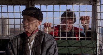 Movie still from “Remo Williams: The Adventure Begins” (1985), directed by Guy Hamilton – Two men are behind bars in a jail cell; Medium shot, Low angle