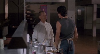 Movie still from “Remo Williams: The Adventure Begins” (1985), directed by Guy Hamilton – A man standing next to another man in a kitchen; Medium shot, Over the shoulder angle