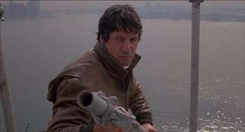 Movie still from “Remo Williams: The Adventure Begins” (1985), directed by Guy Hamilton – A man in a brown leather jacket holding a gun; Close Up shot, Low angle