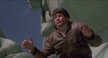 Movie still from “Remo Williams: The Adventure Begins” (1985), directed by Guy Hamilton – A man in a brown jacket is sitting in front of the statue of liberty; Medium shot, Low angle