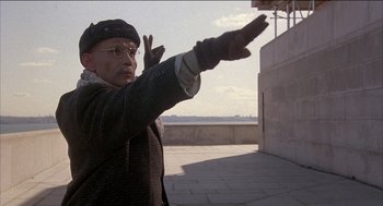 Movie still from “Remo Williams: The Adventure Begins” (1985), directed by Guy Hamilton – An older man in glasses and a hat is holding out his hands; Medium shot, Low angle
