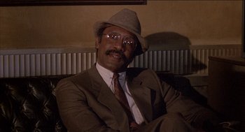 Movie still from “Remo Williams: The Adventure Begins” (1985), directed by Guy Hamilton – A man in a suit and a hat sitting on a couch; Close Up shot, Low angle