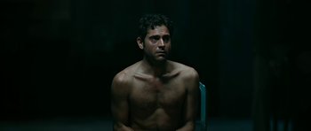 Movie still from “Rendition” (2007), directed by Gavin Hood – A shirtless man sitting in a chair in a dark room; Medium shot, Low angle