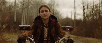 Movie still from “Rhymes for Young Ghouls” (2013), directed by Jeff Barnaby – A woman riding a bike on top of a road; Medium shot, Over the shoulder angle