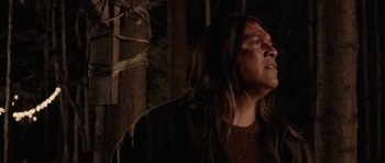 Movie still from “Rhymes for Young Ghouls” (2013), directed by Jeff Barnaby – A man with long hair is standing in a dark room; Medium shot, Low angle