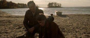 Movie still from “Rhymes for Young Ghouls” (2013), directed by Jeff Barnaby – A man kneeling down next to a woman on a beach; Medium shot, Over the shoulder angle