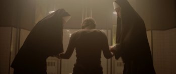 Movie still from “Rhymes for Young Ghouls” (2013), directed by Jeff Barnaby – Two nuns holding hands with a woman in a dark room; Wide shot, Low angle