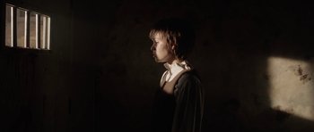 Movie still from “Rhymes for Young Ghouls” (2013), directed by Jeff Barnaby – A young boy standing in a dark room with his head turned to the side; Medium shot, Low angle