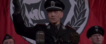 Movie still from “Richard III” (1995), directed by Richard Loncraine – An older man in a military uniform is sitting at a microphone; Close Up shot, Low angle