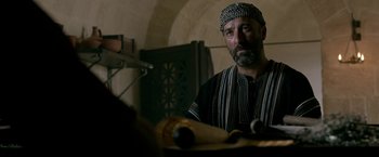 Movie still from “Risen” (2016), directed by Kevin Reynolds – A man wearing a hat is sitting at a table; Medium shot, Over the shoulder angle