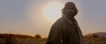 Movie still from “Risen” (2016), directed by Kevin Reynolds – A man with a long beard and a hat is standing in a field with the sun in the background; Medium shot, Low angle
