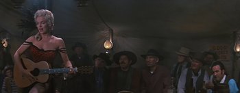 Movie still from “River of No Return” (1954), directed by Otto Preminger – A group of men standing under a tarp with a guitar; Wide shot, Low angle