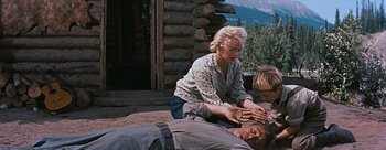 Movie still from “River of No Return” (1954), directed by Otto Preminger – A woman helping a man lay on the ground in front of a log cabin; Medium shot, Low angle