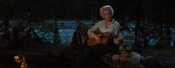 Movie still from “River of No Return” (1954), directed by Otto Preminger – A woman sitting on a log playing a guitar; Medium shot, Low angle