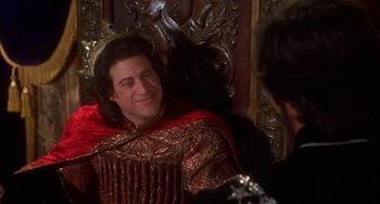 Movie still from “Robin Hood: Men in Tights” (1993), directed by Mel Brooks – A man sitting on top of a chair wearing a crown; Medium shot, Over the shoulder angle