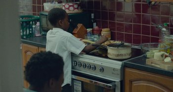 Movie still from “Rocks” (2019), directed by Sarah Gavron – A young boy in a kitchen preparing food; Medium shot, Over the shoulder angle