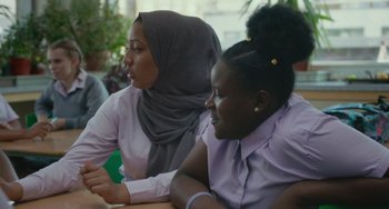 Movie still from “Rocks” (2019), directed by Sarah Gavron – Two women sitting at a table looking at a cell phone; Medium shot, Over the shoulder angle