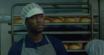 Movie still from “Rocks” (2019), directed by Sarah Gavron – A man wearing an apron standing in front of a rack of baked goods; Close Up shot, Low angle