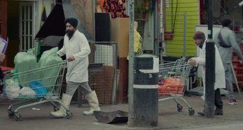 Movie still from “Rocks” (2019), directed by Sarah Gavron – A man in a white lab coat pushing a shopping cart down a street; Wide shot, High angle