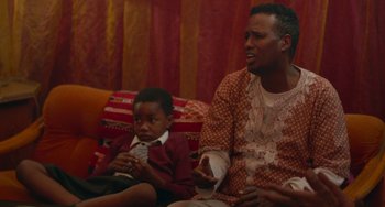 Movie still from “Rocks” (2019), directed by Sarah Gavron – A man and a boy are sitting on a couch; Medium shot, Over the shoulder angle