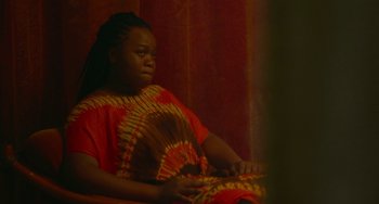 Movie still from “Rocks” (2019), directed by Sarah Gavron – A woman sitting on a chair in a room; Medium shot, Over the shoulder angle