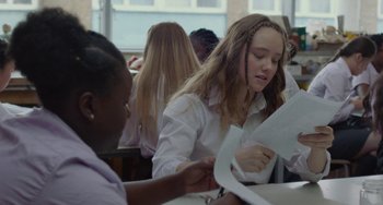 Movie still from “Rocks” (2019), directed by Sarah Gavron – A girl is sitting at a table with papers in her hands; Medium shot, Over the shoulder angle