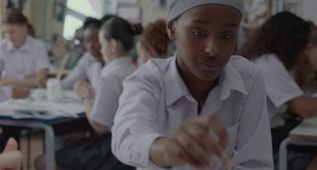 Movie still from “Rocks” (2019), directed by Sarah Gavron – A group of people sitting in a row writing on a piece of paper; Close Up shot, Low angle