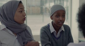 Movie still from “Rocks” (2019), directed by Sarah Gavron – Two young people are sitting together in a room; Medium shot, Over the shoulder angle