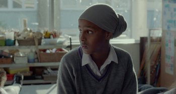 Movie still from “Rocks” (2019), directed by Sarah Gavron – A person sitting in front of a window wearing a hat; Close Up shot, Over the shoulder angle
