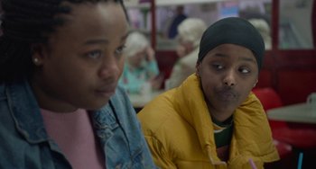 Movie still from “Rocks” (2019), directed by Sarah Gavron – Two young people sitting in a room with people in the background; Close Up shot, Over the shoulder angle