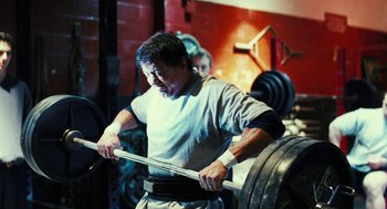 Movie still from “Rocky Balboa” (2006), directed by Sylvester Stallone – A man is lifting a barbell in a gym; Medium shot, Over the shoulder angle