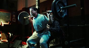 Movie still from “Rocky Balboa” (2006), directed by Sylvester Stallone – A man lifting a barbell while another man watches; Medium shot, Low angle