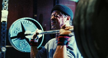 Movie still from “Rocky Balboa” (2006), directed by Sylvester Stallone – A man holding a barbell while wearing a hat; Close Up shot, Low angle
