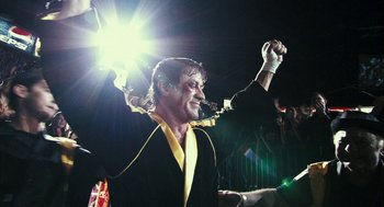 Movie still from “Rocky Balboa” (2006), directed by Sylvester Stallone – A man in a black and yellow outfit holding his hands up; Medium shot, Low angle