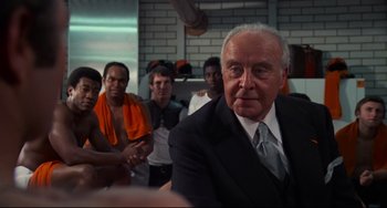 Movie still from “Rollerball” (1975), directed by Norman Jewison – An older man in a suit and tie with a group of people; Medium shot, Low angle